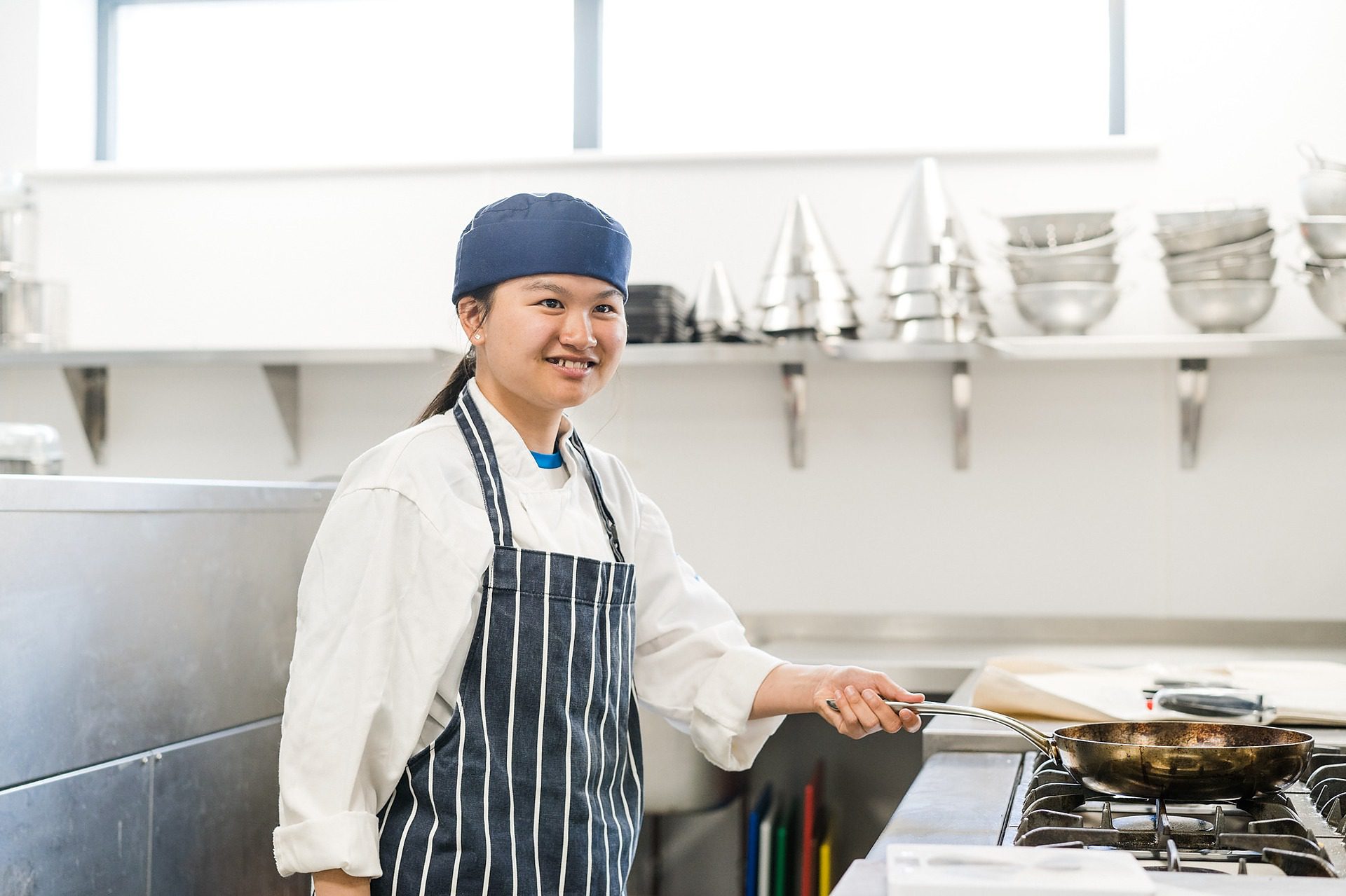 Student wearing blue apron and a toque standing preparing food in kitchen