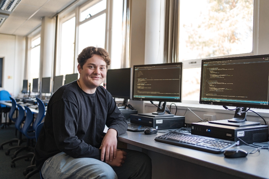 Male student in black jumper sitting in front of a row of computer desktops smiling