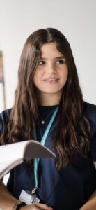 Student with long hair wearing a Harrogate College lanyard, smiling and looking to the left, behind an open textbook being held by someone else not in the image