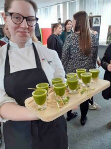 Female wearing apron serving green juices on a wooden tray at Sustainability Show with a group of people at the background