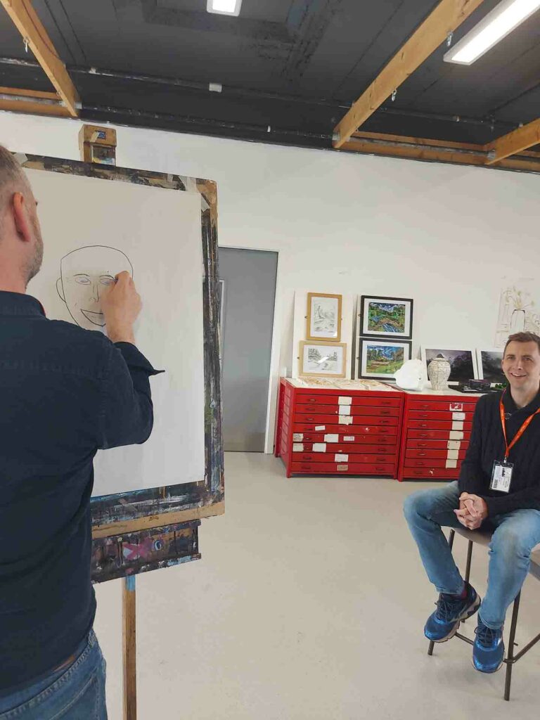 A man drawing a face of a man on the drawing stand board, the model man sitting down smiling in art classroom