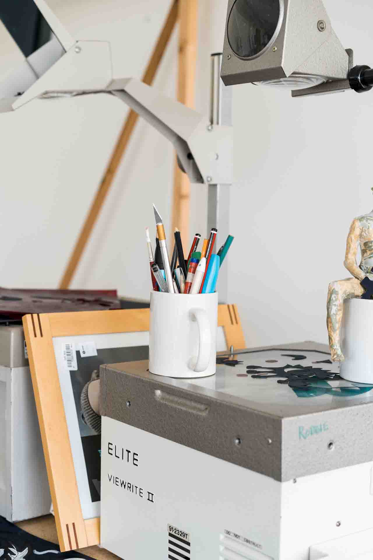 A white mug full of pencils, pens and markers on top of the Elite Viewrite Overhead Projector in the art classroom