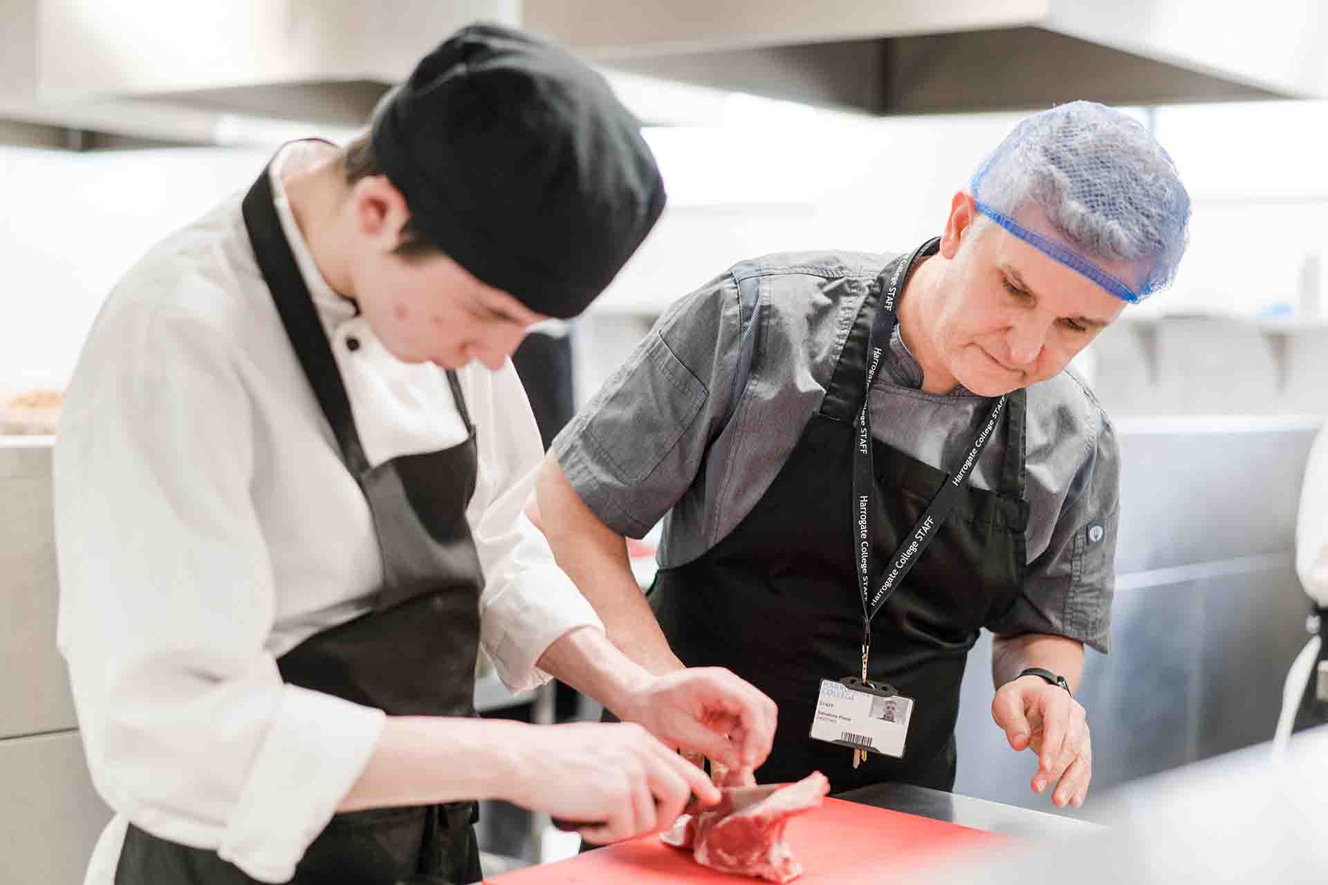 Mature catering supervisor in grey shirt, wearing hair net and black apron assisting a male student wearing kitchen uniform and black apron, in cutting a piece of meat in the kitchen