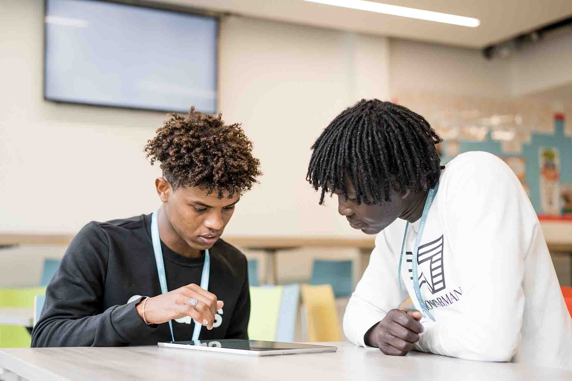 Two African male students both wearing Harrogate College lanyard sitting in classroom looking at an iPad