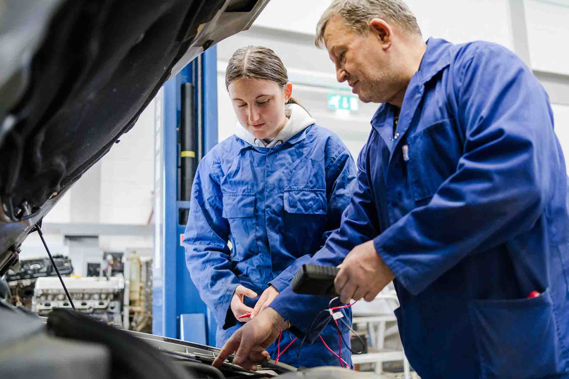 Mature motor vehicle lecturer in blue uniform with a probe circuit tester in hand showing the engine of a car to a female student in blue uniform standing with the car bonnet cover open