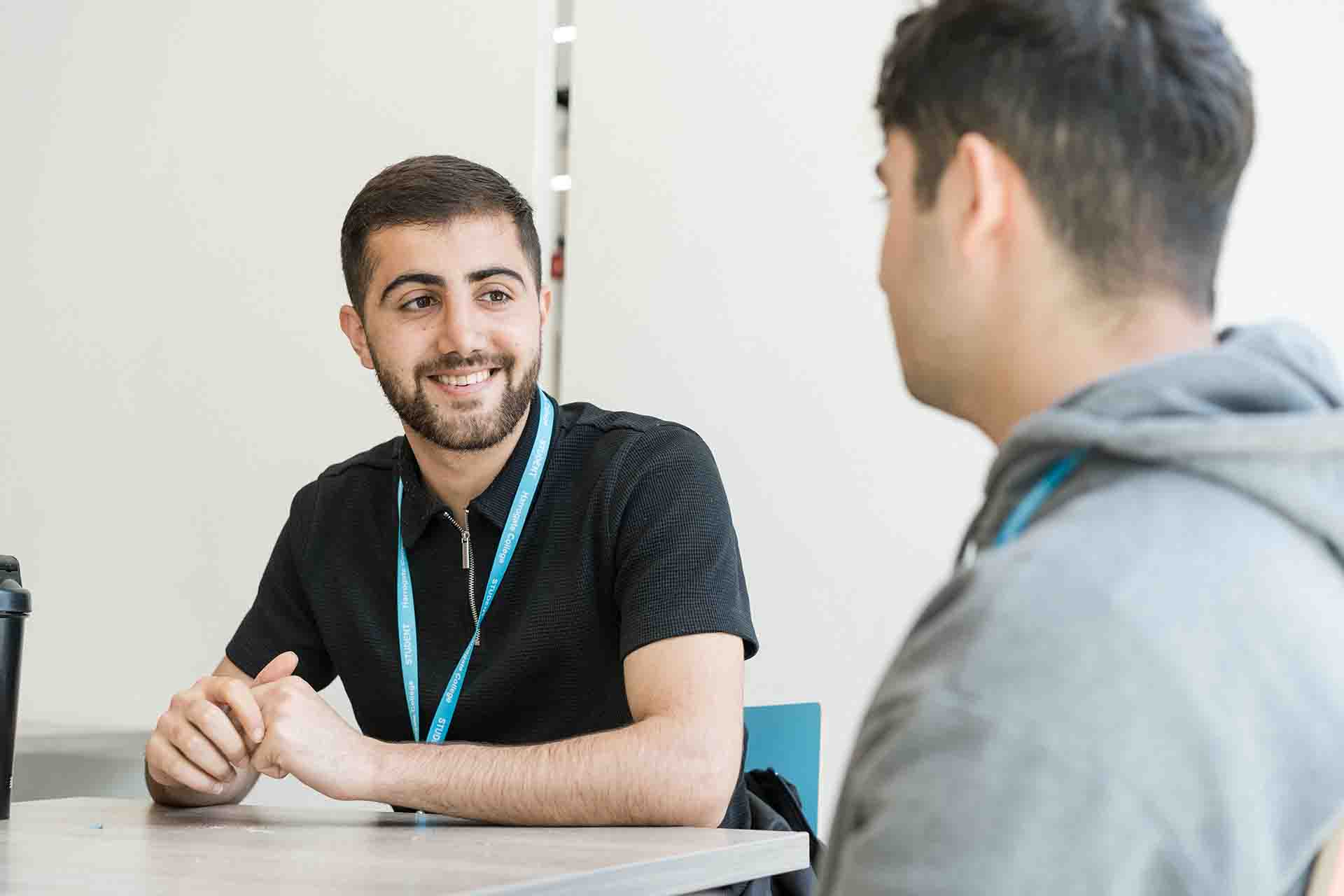 A male student wearing black top and Harrogate College lanyard sitting on a table smiling looking to his left to another male student