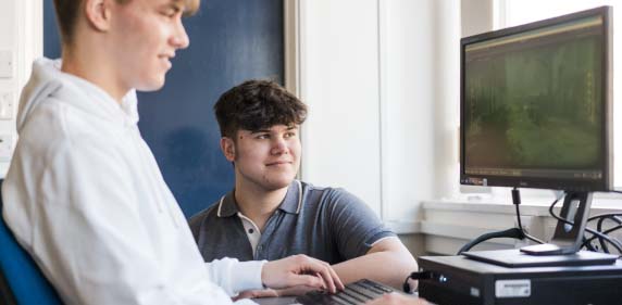 Two students sat looking at a desktop computer together smiling whilst working