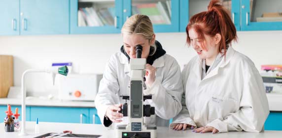 Two students wearing lab coats in the science lab looking into a microscope