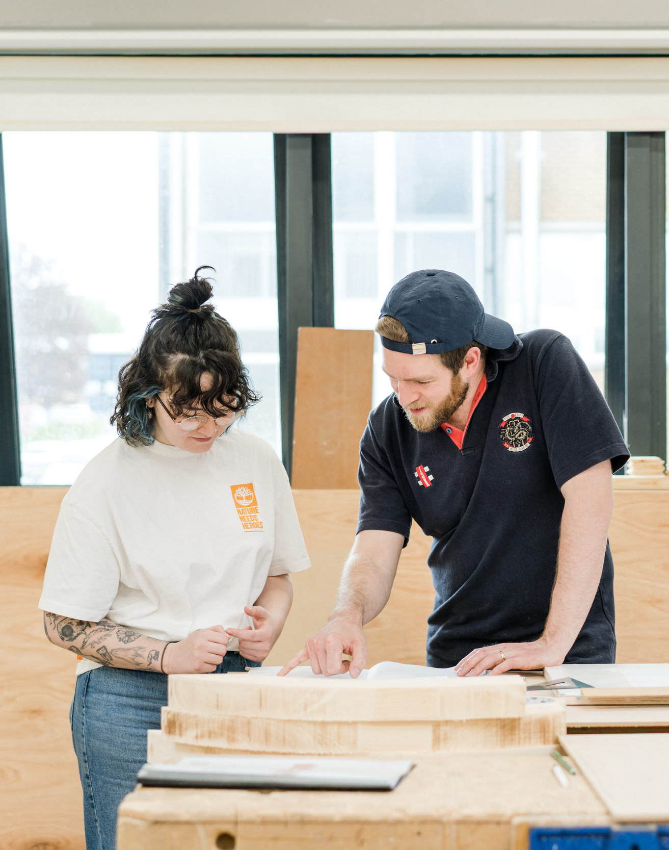 Student wearing a cap backwards, standing in the carpentry workshop pointing the paperwork and explaining to another student in white T shirt