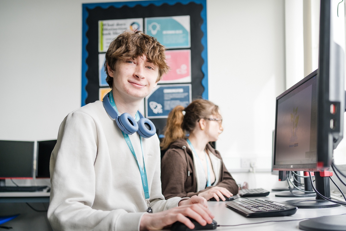 Student wearing blue lanyard with headphone sitting in front of computer smiling