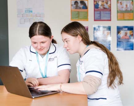 Two Harrogate College students wearing white college overalls sat at a desk in a classroom working on a laptop together smiling