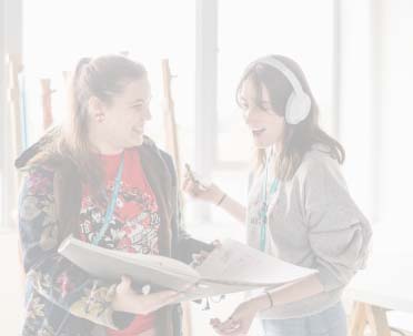 Two art students looking down at coursework and smiling, one wearing headphones