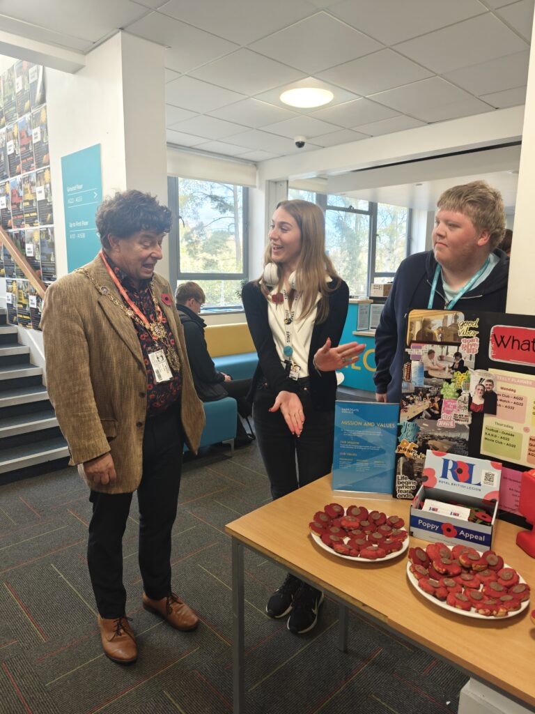 two men stand with a woman by a table containing remembrance day displays and plates of poppy shaped biscuits