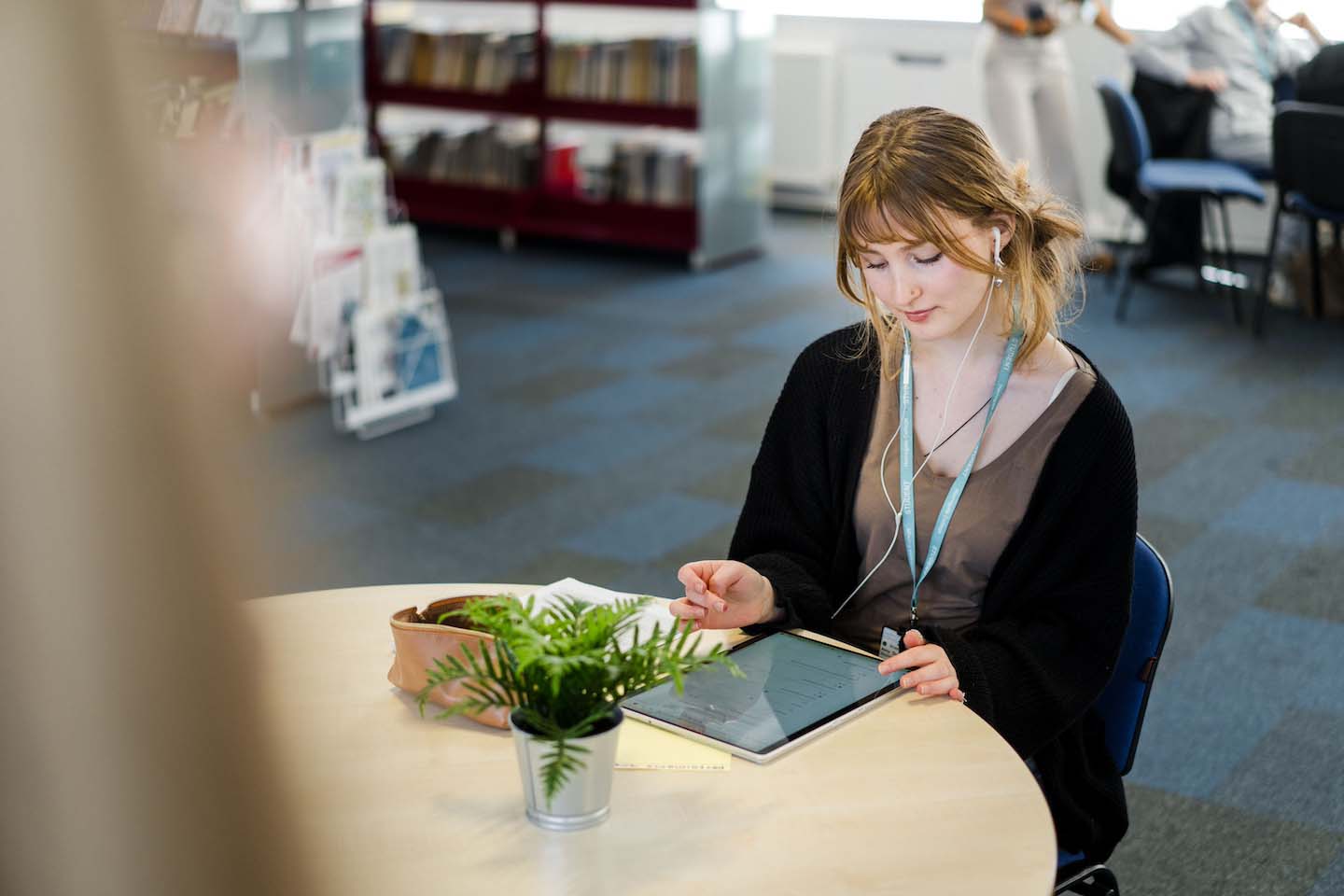 Harrogate College student sat in the library wearing lanyard and headphones looking down at a tablet