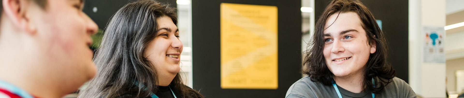 Three students sat in Harrogate College smiling and chatting wearing Harrogate College blue lanyards