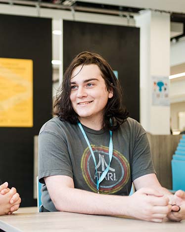 Harrogate College student sat in the college wearing a lanyard and smiling at a friend sat out of shot