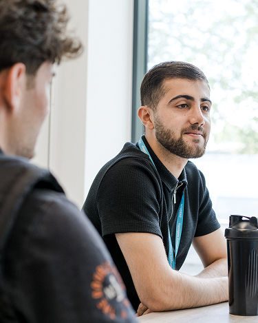 Harrogate College student sat in the college wearing a lanyard and chatting to a friend sat out of shot