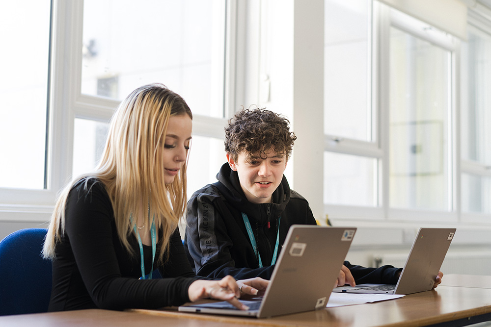 Two students sat in classroom with laptops on desk looking at work on laptop screen