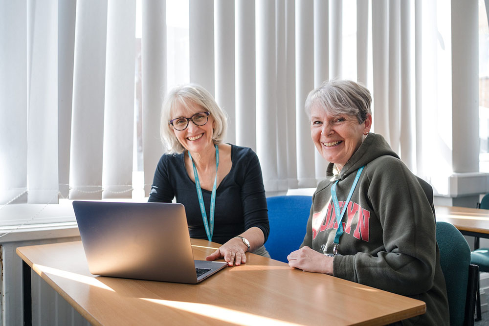 Two adults sat at a desk in front of a laptop smiling at a camera
