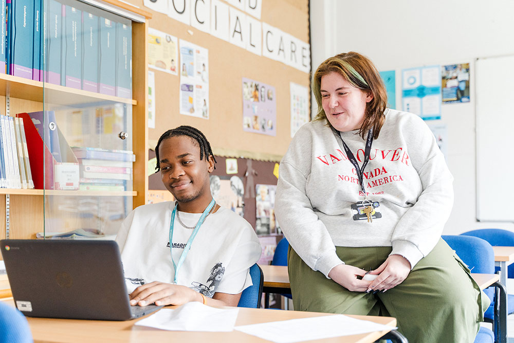 Student sat at desk in Harrogate College classroom next to teacher both looking at laptop screen