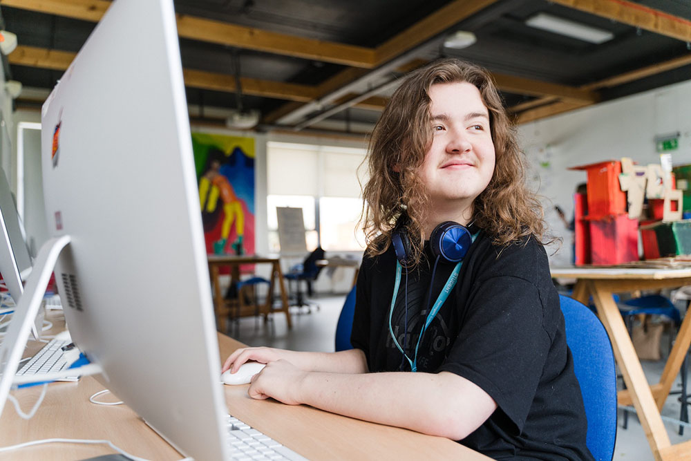 Harrogate College student sat in front of an apple mac desktop computer with headphones around neck, looking to the side and smiling