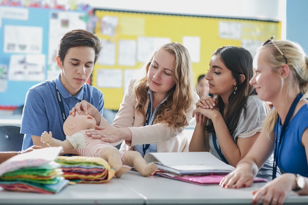 Students being shown by teacher how to do first aid on a baby mannequin