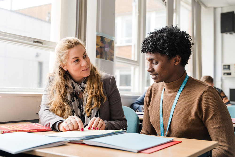 Teacher and student sat at a desk in Harrogate College classroom, teacher pointing and explaining something in a textbook