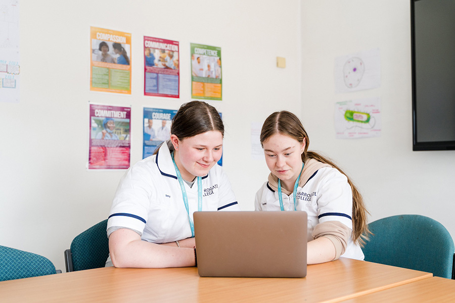 Two Health students in medical nurse uniforms sat in a classroom looking at a laptop together