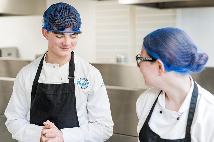 Two catering students wearing kitchen overalls, aprons and hairnets chatting in the kitchen