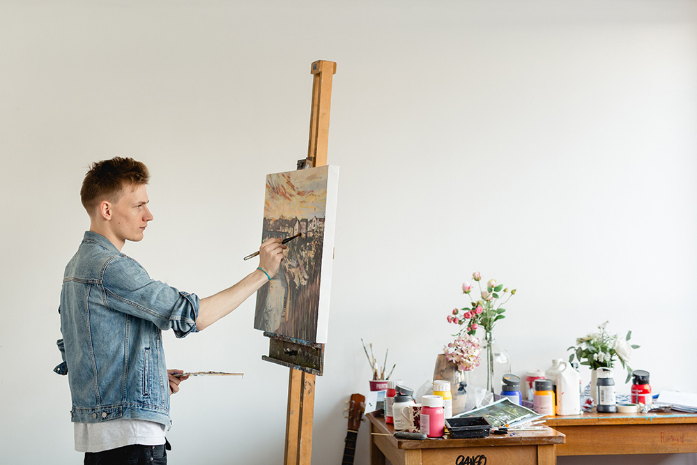 Student stood painting canvas on an easel in the art studio at Harrogate College next to a table of paints and flowers