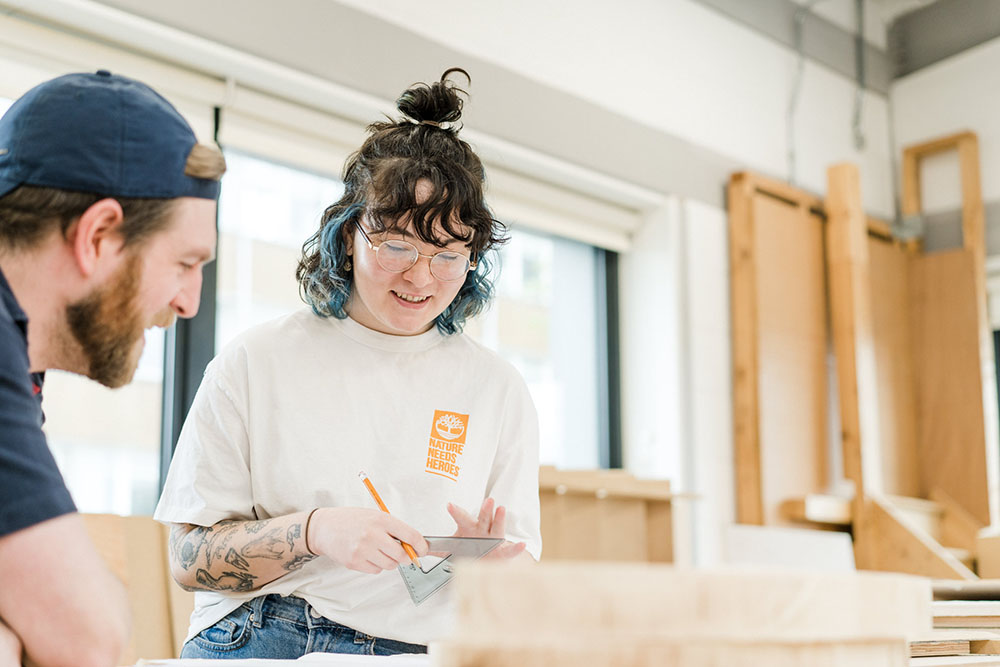 Student and teacher in wood workshop looking down measuring wood, with wooden boards in background