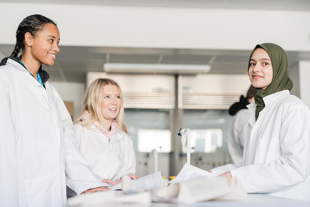 Three students in stood in lab coats in the Science laboratory at Harrogate College smiling