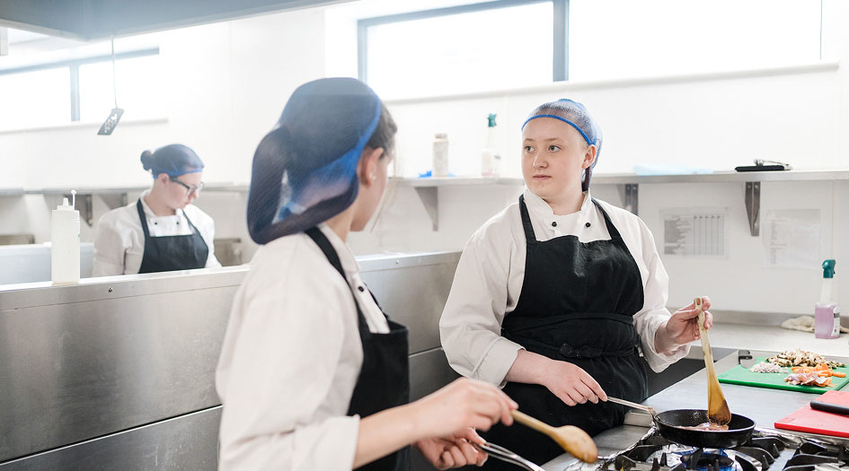 Catering students wearing kitchen overalls, aprons and hairnets cooking in the kitchen