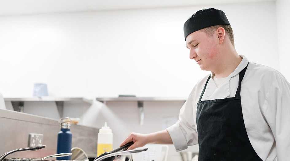 Catering student wearing kitchen overalls, aprons and hat cooking in the kitchen