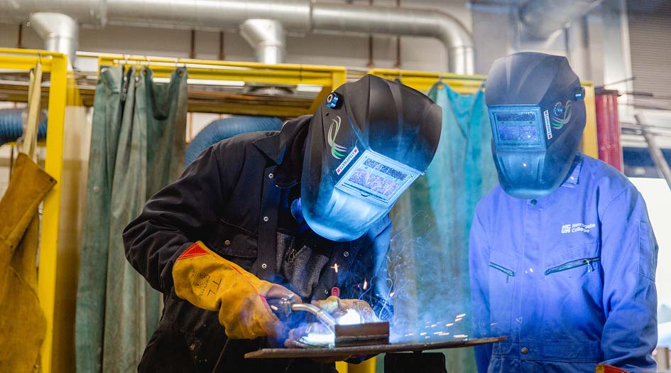 Mechanical fabrication students wearing welding helmet on-site using machinery for cutting, bending, welding, and assembling metal