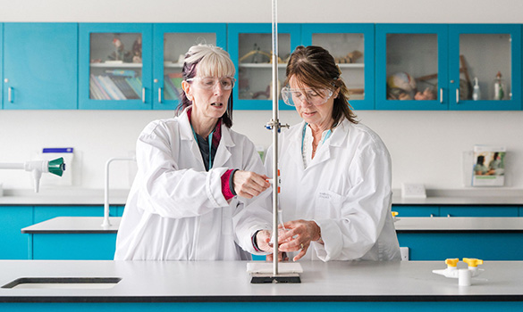 Two adult learners in Science classroom, wearing lab coats and safety goggles using a bunsen burner