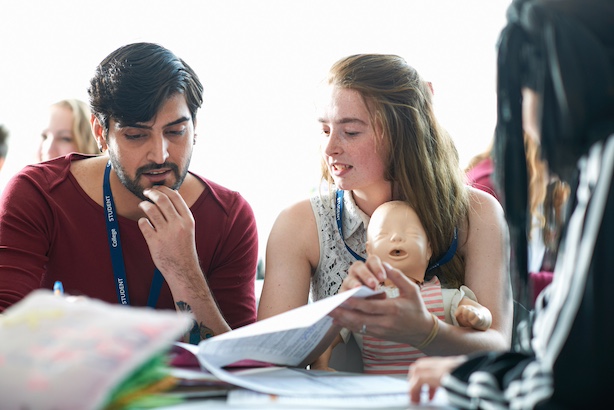 Two students sat with a dummy baby looking at paperwork and practising holding the baby