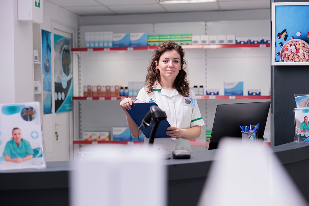 Student stood in pharmacy smiling at camera holding a clipboard