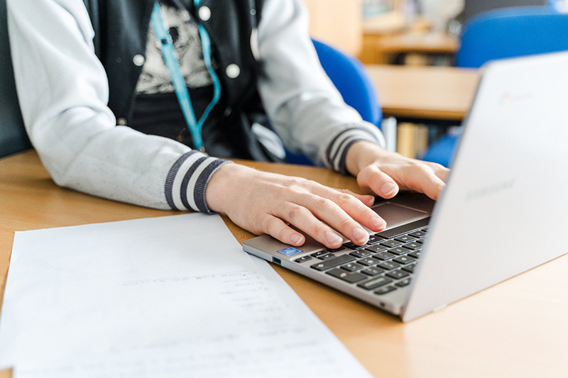 Student's hands typing on a laptop in a classroom wearing Harrogate College lanyard