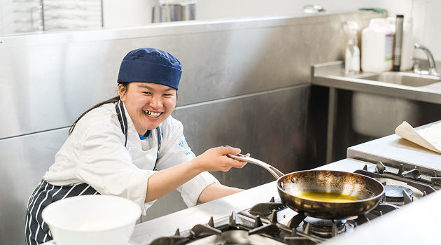 Student in kitchen wearing chef overalls turning the hob on whilst holding a frying pan