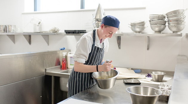 Student in kitchen wearing chef overalls whisking a mixture together in a bowl and smiling