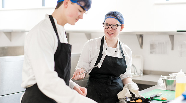 Two students in chef overalls and hair nets chatting whilst cooking over a hob in training kitchen