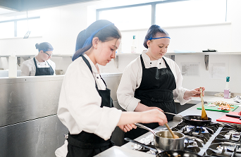 Two students in chef overalls and hair nets chatting whilst cooking over a hob in training kitchen