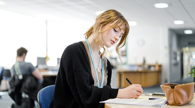 Student sat at desk looking down and writing notes in study book