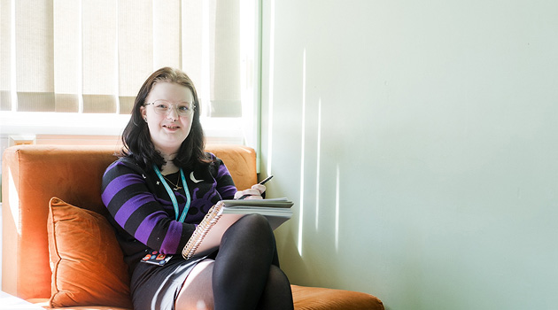 Student sat on sofa working on learning books and a pen on her lap, smiling at camera