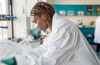 Student in lab overalls leaning over and focusing on an experiment in the Science Lab surrounded by lab equipment