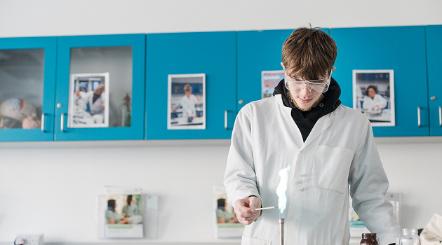 Student in Science Laboratory wearing lab overalls and goggles lighting a bunsen burner