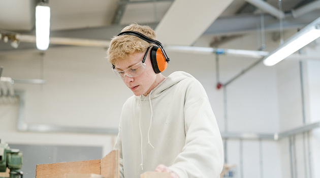 Student stood in workshop looking down at woodwork project whilst wearing safety goggles and ear defenders