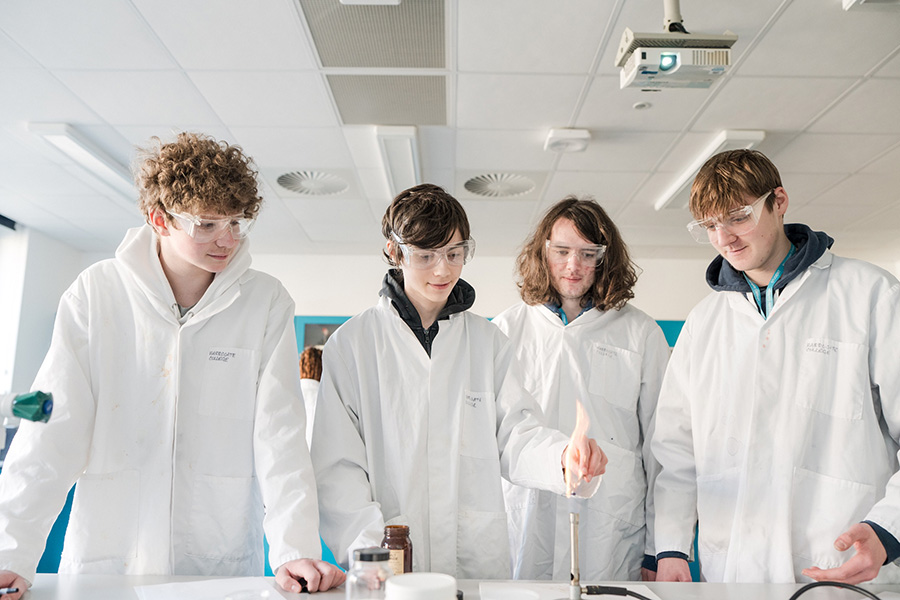 Four students stood in the Science laboratory wearing lab coats and goggles, watching one student light a bunsen burner