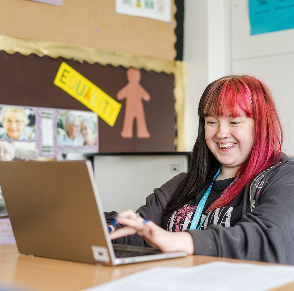 Student sat at desk typing on laptop smiling and looking happy
